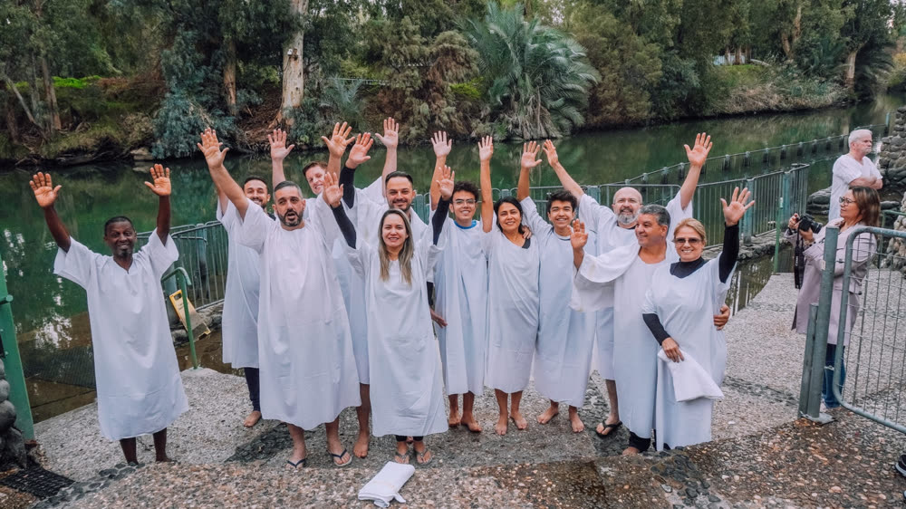 Grupo de túnicas brancas com as mãos levantadas em celebração diante do Rio Jordão.